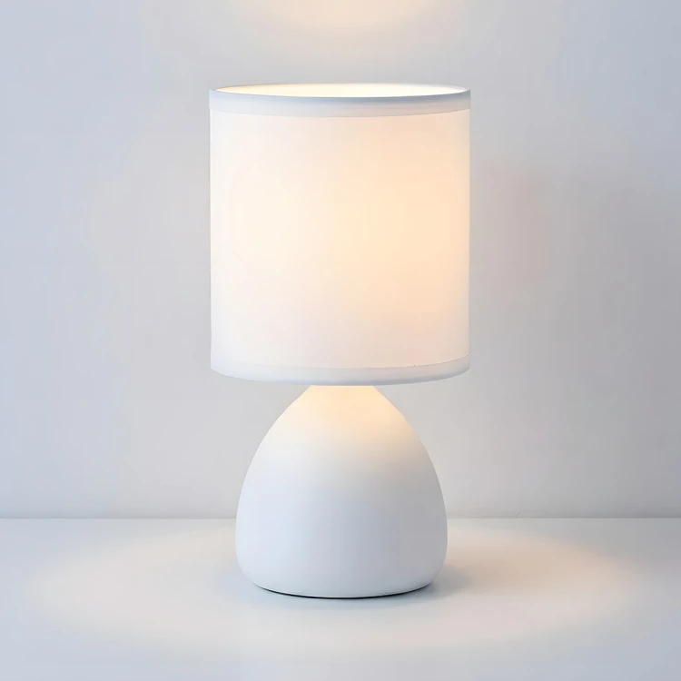 Lampka nocna SABRINA 1×E14 doskonale sprawdzi się na stoliku przy łóżku, komodzie lub biurku. Biała ceramika i tkaninowy abażur pasują do jasnych, nowoczesnych wnętrz. Wysokość lampki wynosi 23,5 cm. Żarówka nie jest dołączona.